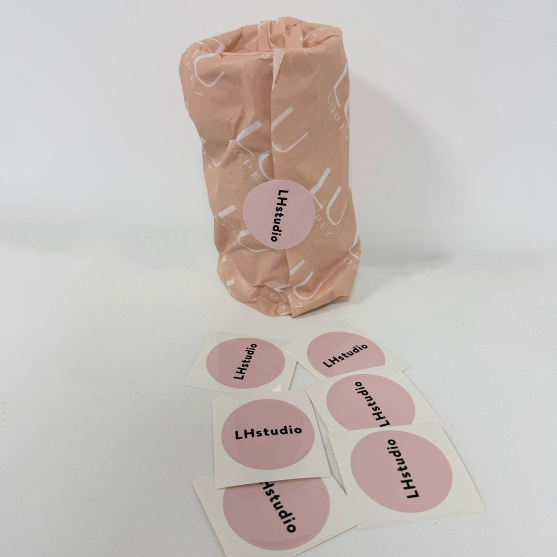 Round pink coated paper stickers with a studio logo beside a kraft-wrapped product, glossy coated finish