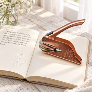 An open book on a sunlit wooden table with an embossed mark on the page and a rose gold embosser resting beside it