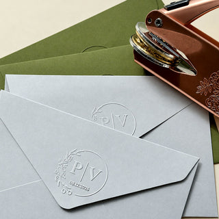 A grey envelope flap embossed with a crisp monogram seal, with olive envelopes and a rose gold embosser behind