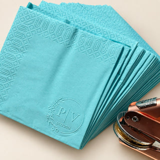 A fan of turquoise paper napkins embossed with a monogram seal, beside a rose gold embosser