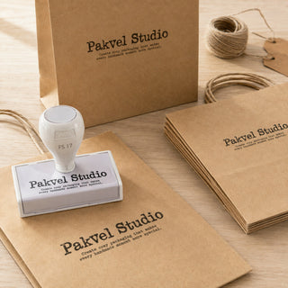 Custom pre-inked stamp on a wooden table beside kraft paper bags stamped with a brand name and tagline, with twine and hang tags