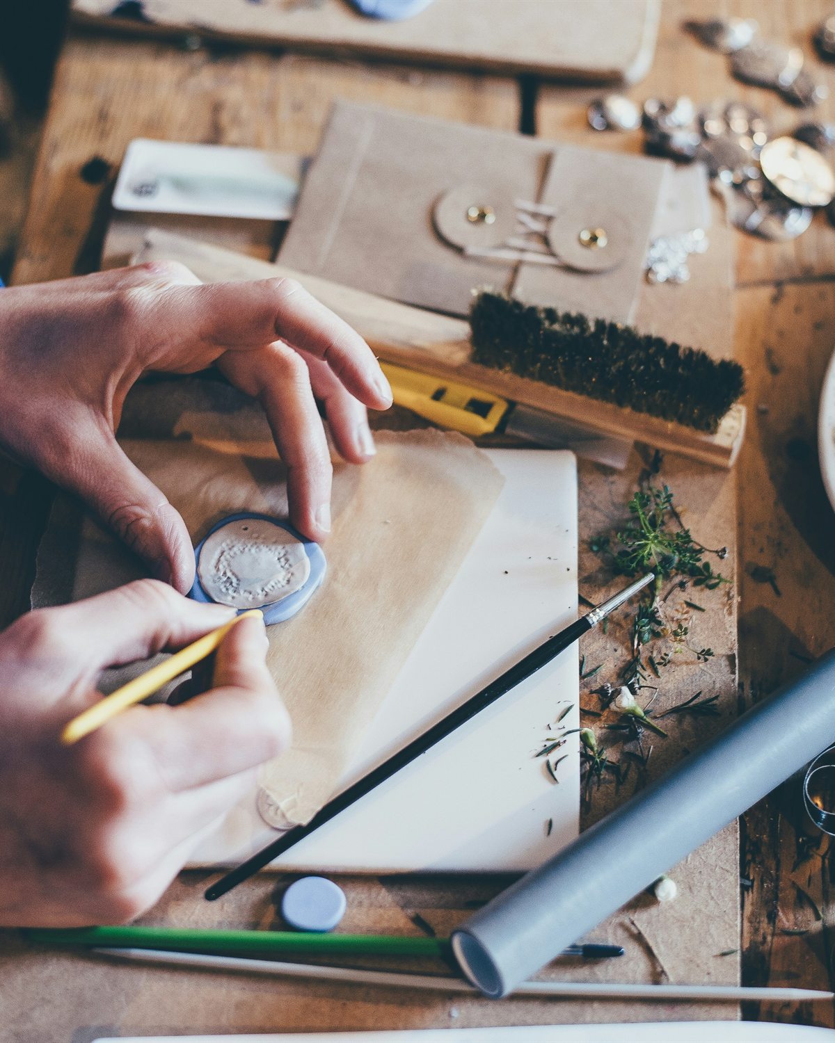 Hands at a maker workbench crafting wax seals among kraft paper, tools and dried flowers